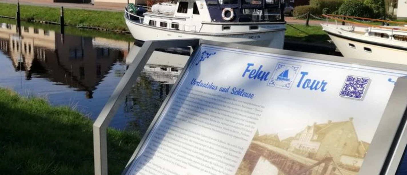 Fehn-Tour.png Infotafel zum historischen Rundgang vor einem Kanal mit Boot und Häusern in Rhauderfehn