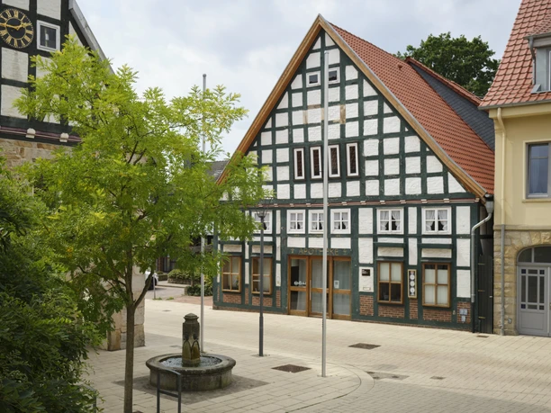 Fachwerkgebäude säumen einen kleinen, gepflasterten Platz mit einem dekorativen Brunnen im Vordergrund.