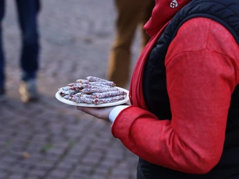 VIP_Fuehrung (5).JPG Person in roter Jacke hält Teller mit kleinen Wurstwaren auf einem gepflasterten Platz.