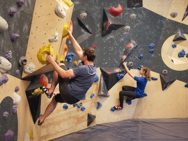 Two people are climbing on an indoor climbing wall with colorful holds; the man is wearing a gray shirt, the woman a blue one.