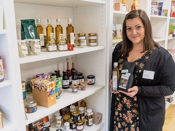 On y voit Mme Uhlmann, debout devant une armoire remplie de produits régionaux de la région, dans l'office de tourisme de Rochlitzer Muldental.
