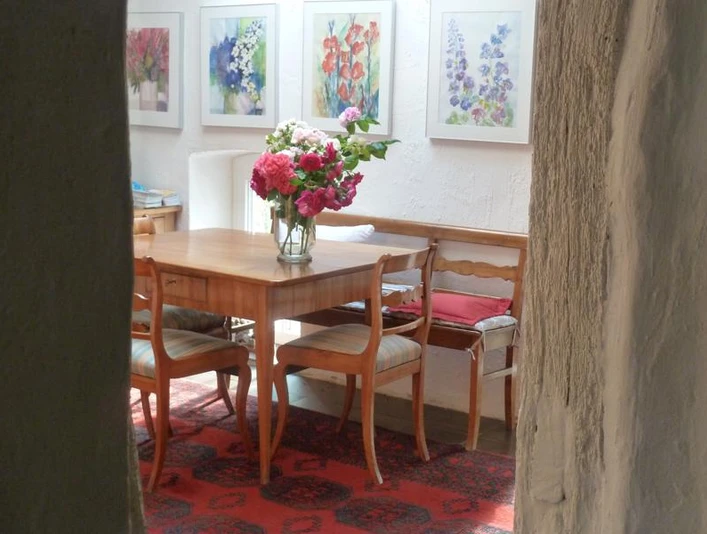 P1070270-800x600_Bei-Veronica.jpg Ein heller Raum mit Holzmöbeln, Blumenstrauß auf dem Tisch, umgeben von farbenfrohen Gemälden.