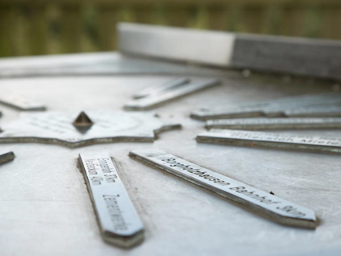 Stadt_Borgholzhausen_Urheber-immer-mit-angeben-direkt-am-Bild (12).jpg Metallplatte mit Richtungsschildern auf einem Aussichtspunkt, zeigt Entfernungen zu nahegelegenen Zielen.