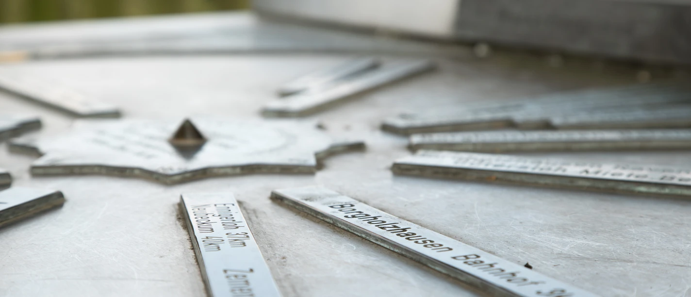 Stadt_Borgholzhausen_Urheber-immer-mit-angeben-direkt-am-Bild (12).jpg Metallplatte mit Richtungsschildern auf einem Aussichtspunkt, zeigt Entfernungen zu nahegelegenen Zielen.