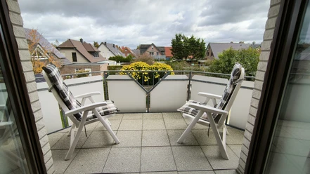Balkon.jpg Zwei weiße Gartenstühle auf einem Balkon mit Blumenkasten, umgeben von Wohnhäusern und bedecktem Himmel.