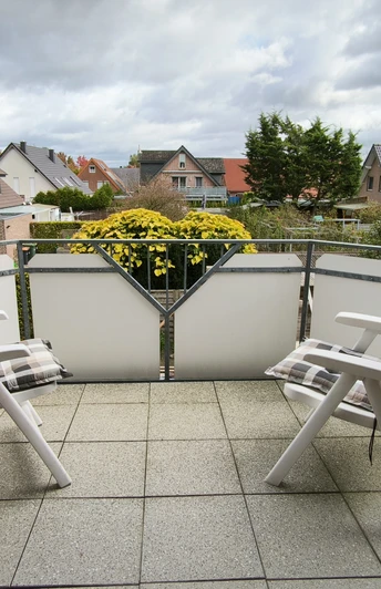 Balkon.jpg Zwei weiße Gartenstühle auf einem Balkon mit Blumenkasten, umgeben von Wohnhäusern und bedecktem Himmel.