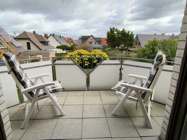 Balkon.jpg Zwei weiße Gartenstühle auf einem Balkon mit Blumenkasten, umgeben von Wohnhäusern und bedecktem Himmel.