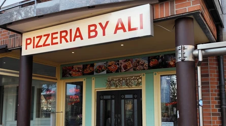 Pizzeria_by_Ali_Spelle©SG-Spelle.jpg Eingang der Pizzeria By Ali mit beleuchtetem Schild und Fotos von Gerichten über der Tür.