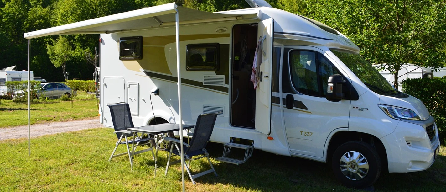 Wohnmobil mit ausgefahrener Markise steht auf grünem Rasen; Campingstühle und Tisch davor, umgeben von Bäumen.