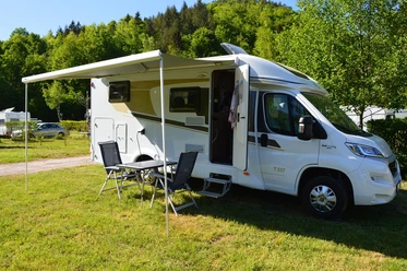 Pixabay_Caravanning-3509593_1280_Copyright_Reipen.jpg Wohnmobil mit ausgefahrener Markise steht auf grünem Rasen; Campingstühle und Tisch davor, umgeben von Bäumen.