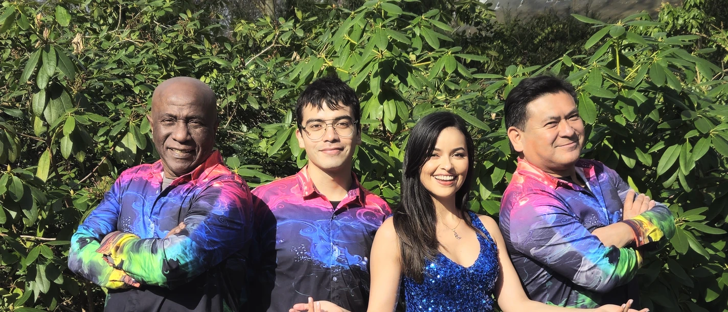 Foto 3 Sal Y Mango.jpg Vier mensen staan glimlachend naast elkaar voor een groen bladerdak. Twee mannen dragen kleurrijke shirts, een vrouw een glinsterende blauwe jurk en een andere man een zwart shirt met kleurrijke accenten. Ze zien er allemaal gelukkig en vrolijk uit.