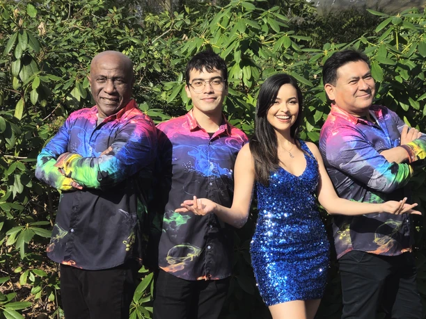 Foto 3 Sal Y Mango.jpg Vier Menschen stehen lächelnd nebeneinander vor grünem Laub. Zwei Männer tragen bunte Hemden, eine Frau ein glitzerndes, blaues Kleid und ein weiterer Mann ein schwarzes Hemd mit bunten Akzenten. Alle wirken zufrieden und fröhlich.