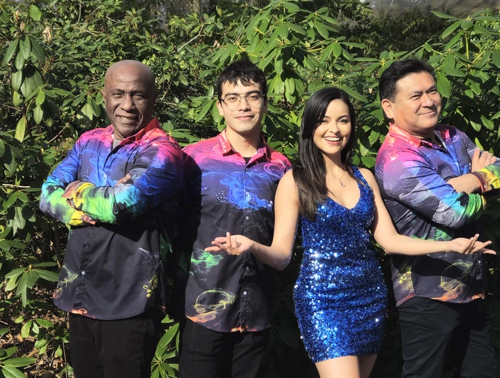 Foto 3 Sal Y Mango.jpg Vier Menschen stehen lächelnd nebeneinander vor grünem Laub. Zwei Männer tragen bunte Hemden, eine Frau ein glitzerndes, blaues Kleid und ein weiterer Mann ein schwarzes Hemd mit bunten Akzenten. Alle wirken zufrieden und fröhlich.