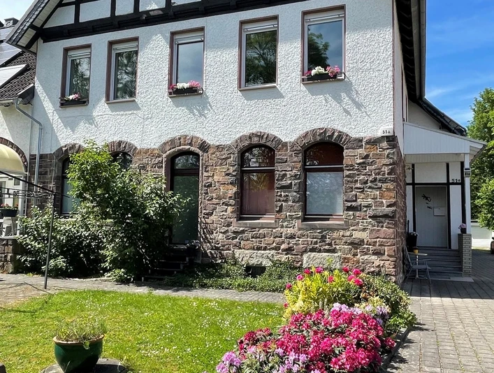 Zweistöckiges Fachwerkhaus mit Steinbasis, blühenden Beeten und gepflegter Rasenfläche im Vordergrund.