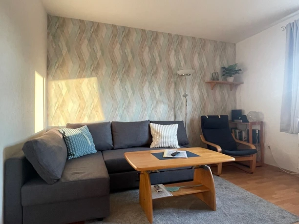 Wohnzimmer mit grauem Ecksofa, Holztisch, gemusterter Tapete, Teppich und moderner Lampe.