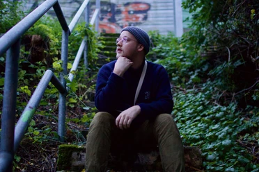 Eine Person mit Mütze sitzt nachdenklich auf einer Treppe inmitten grüner Vegetation.