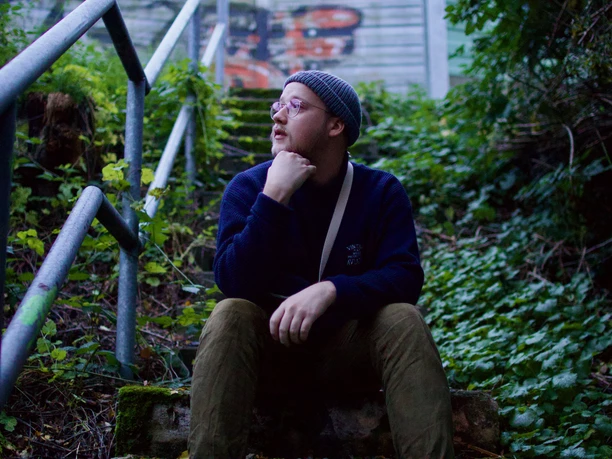 Eine Person mit Mütze sitzt nachdenklich auf einer Treppe inmitten grüner Vegetation.