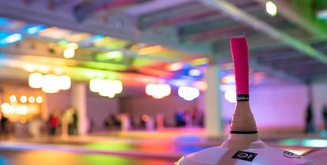 csm_eisspielzeiten-owl_58c81c1c7d.jpg Eine bunte Eishalle mit Besuchern im Hintergrund, im Fokus ein Eisstock mit pinker Griffmanschette.