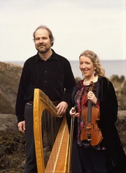 Maire Breatnach -Thomas Loefke Maire Breatnach und Thomas Loefke stehen nebeneinander am Meer. Maire hält eine Geige, Thomas eine Harfe.