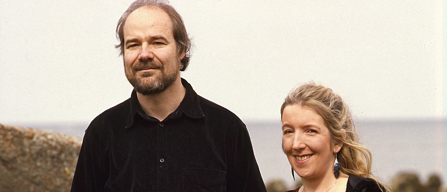 Maire Breatnach -Thomas Loefke Maire Breatnach und Thomas Loefke stehen nebeneinander am Meer. Maire hält eine Geige, Thomas eine Harfe.