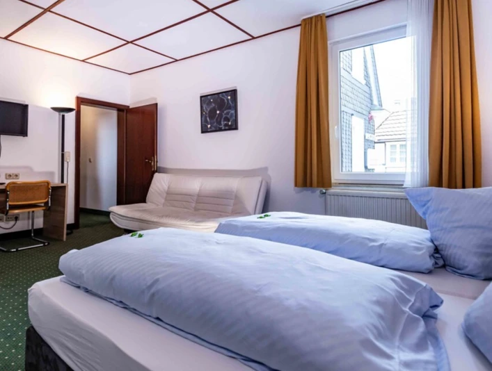 Hotelzimmer im Hotel Kimmeskamp in Velbert Ein helles Hotelzimmer mit einem Doppelbett, einem kleinen Sofa, Schreibtisch und Fenster mit Vorhang.