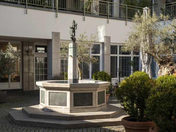 Löwenbrunnen Löwenbrunnen in einem Innenhof mit grünen Pflanzen und einer kleinen Löwenfigur auf der Spitze.