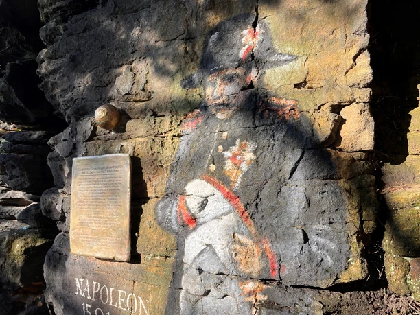 Napolenonstein Bildnis und Kugel Ein farbiges Wandgemälde von Napoleon auf einer Felswand, daneben eine Gedenktafel und eine Kanonenkugel; die Szene ist sonnig beleuchtet.