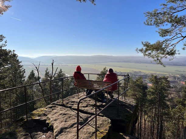 Aussichtspunkt Napoleonstein Zwei Personen in roten Jacken sitzen auf einer Bank auf einem Felsvorsprung, umgeben von Bäumen, mit Blick auf eine weite, sonnige Landschaft.