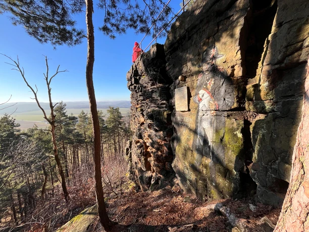 Napoleonstein Person in rotem Mantel steht auf Felsvorsprung mit Blick auf bewaldete Landschaft; Felswand zeigt Napoleon-Graffiti und Gedenktafel.