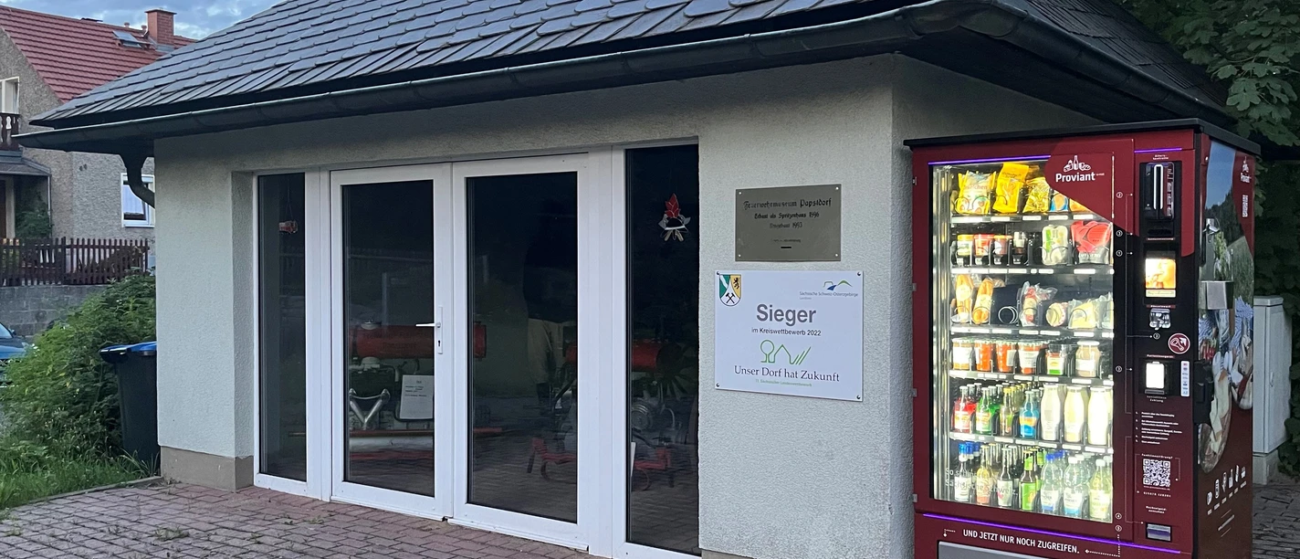 Feuerwehrmuseum Papstdorf mit Proviantomat Kleines Gebäude mit Schieferdach, daneben ein roter Automat mit beleuchteten Flaschen und Snacks.