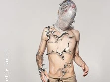 Person mit Netz über dem Kopf und abstrakten Formen am Oberkörper vor hellem Hintergrund