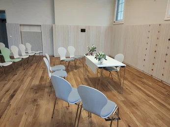 Ein heller Raum mit Holzfußboden, weißen Stühlen und einem zentralen Tisch mit Blumen.