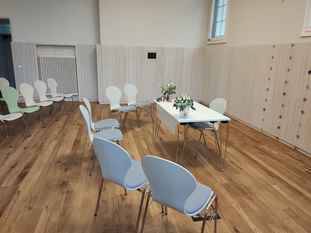 Ein heller Raum mit Holzfußboden, weißen Stühlen und einem zentralen Tisch mit Blumen.