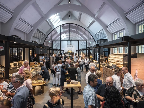 Markthalle_Herford_30.8.19_lowres_167.jpg Menschenmenge in der Markthalle Herford, umgeben von Ständen mit frischen Lebensmitteln und Produkten.
