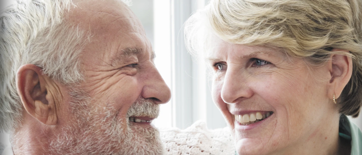 AWO Wohnberatung - Ehepaar.jpg Ein älteres Ehepaar sitzt liebevoll lächelnd nebeneinander, die Frau legt sanft ihre Hand auf den Arm des Mannes.