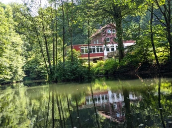 1Teich_An den Teichen Rotes Haus am Waldrand spiegelt sich im ruhigen Teich, umgeben von dichtem grünen Laub.