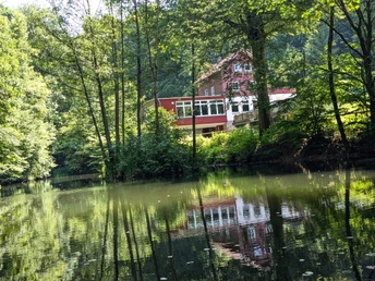 Teich_An den Teichen Rotes Haus am Waldrand spiegelt sich im ruhigen Teich, umgeben von dichtem grünen Laub.