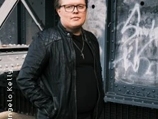 Angelo Kelly steht in lässiger Kleidung vor einer graffiti-besprühten Metallwand und blickt direkt in die Kamera.