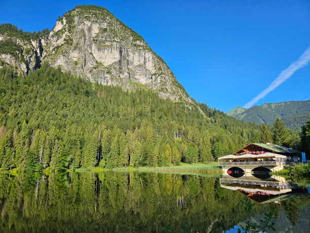 Berggasthof Pflegersee Berggasthof am stillen See vor bewaldetem Hang und steilem Felsmassiv im Sonnenlicht