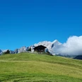 Eckbauer Holzhaus auf grünem Hügel vor schroffen Bergen und einem tiefblauen Himmel mit vorbeiziehender Wolke