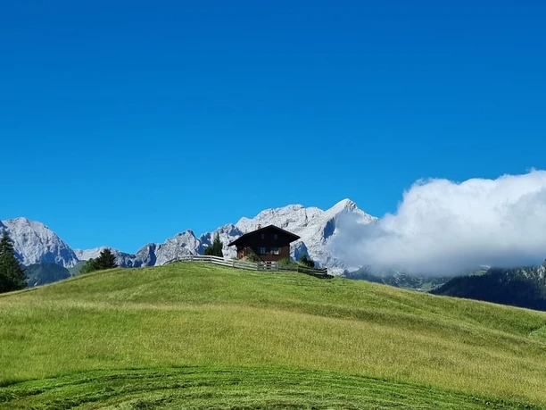 Eckbauer Holzhaus auf grünem Hügel vor schroffen Bergen und einem tiefblauen Himmel mit vorbeiziehender Wolke