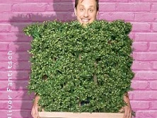 Ein Mensch schaut lächelnd über die Spitze eines rechteckigen, dichten grünen Buschwerks.