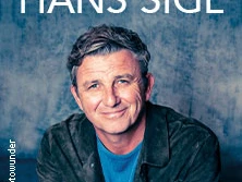 Hans Sigl lächelt freundlich vor einem neutralen Hintergrund in die Kamera, bekleidet mit einer dunklen Jacke.