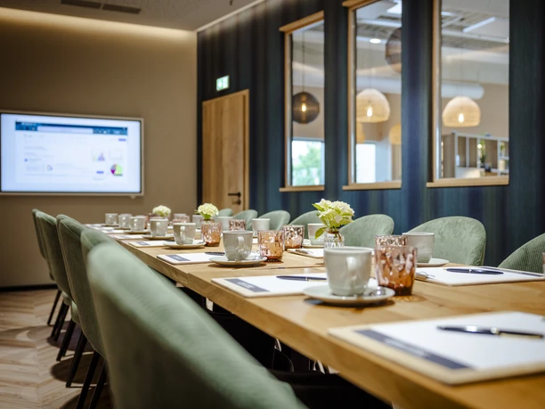 4-Tagungsraum.jpg Ein moderner Konferenzraum mit langer Tafel, grünen Stühlen, Schreibutensilien und Kaffee-Setting.