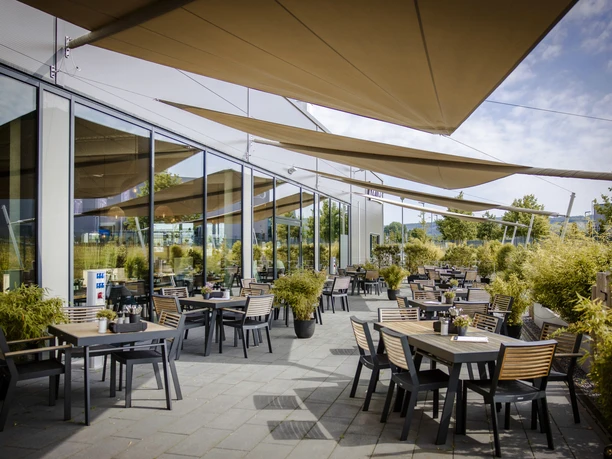 5-Terrasse.jpg Sonnige Terrasse eines modernen Cafés mit schattenspendenden Segeln und stilvollen Sitzgelegenheiten.