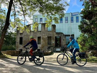 Zwei Radfahrer fahren vor einem modernen Gebäude mit Glasfassade entlang, umgeben von Bäumen.