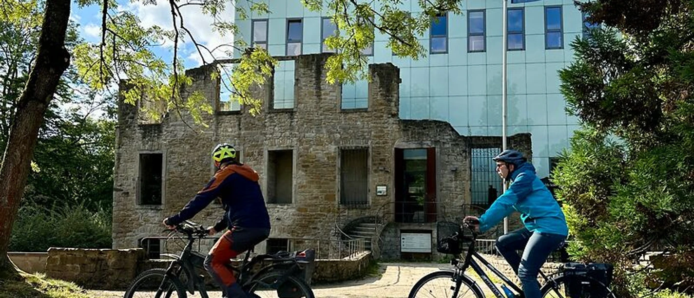 Zwei Radfahrer fahren vor einem modernen Gebäude mit Glasfassade entlang, umgeben von Bäumen.
