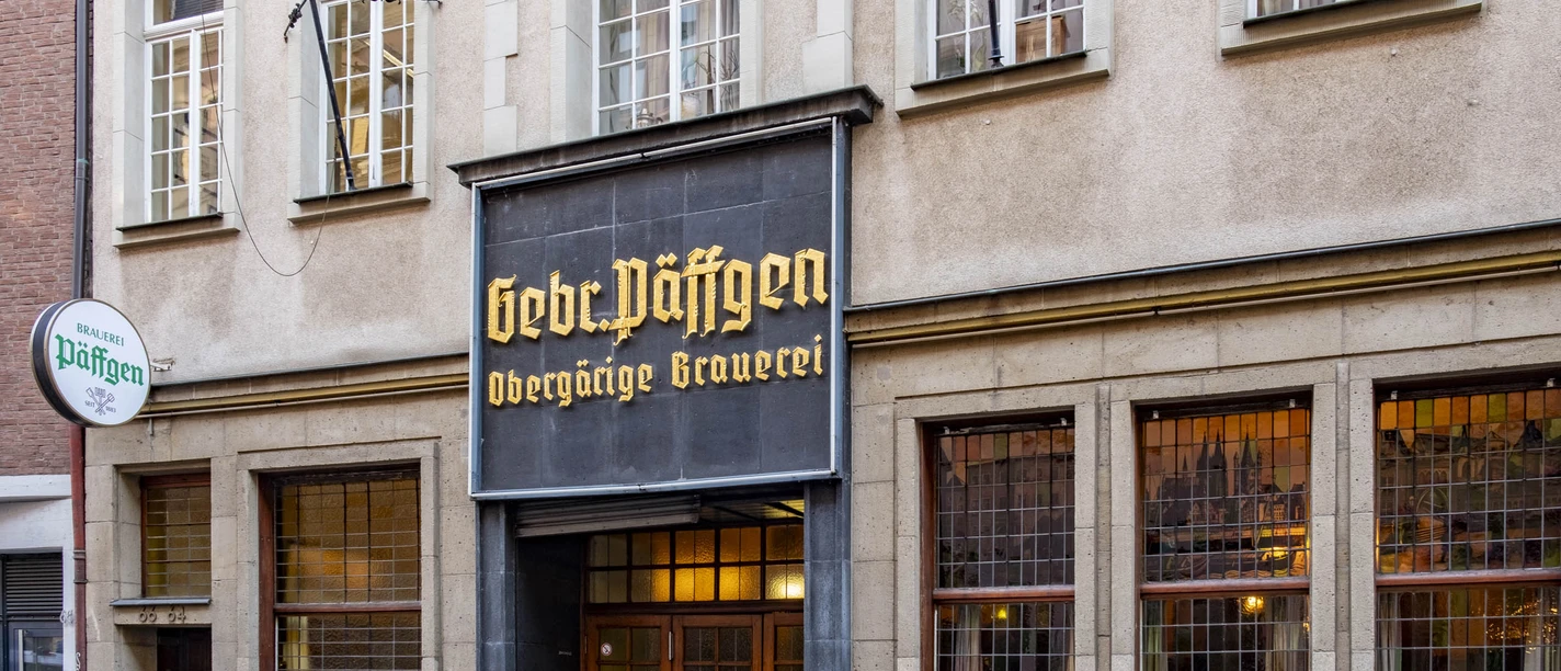 Brauerei Päffgen Eingang zum Brauhaus durch eine rustikale Holztür, darüber ein mit goldenen Buchstaben verziertes Schild.