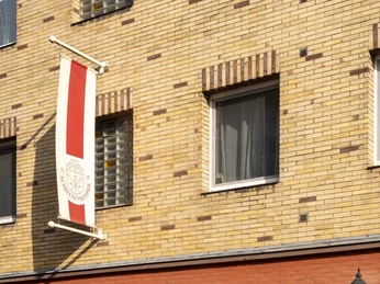 Brauhaus Schreckenskammer Ansicht der Fassade des Brauhauses Schreckenskammer in Köln. Das Gebäude zeigt eine einstöckige rote Backsteinfront mit dem Schriftzug "Biergarten" in einem Fenster und drei Wandlaternen, die an der Fassade befestigt sind. Eine rot-weiße Fahne mit dem Wappen des Brauhauses hängt an der oberen Etage des gelb-rot gemusterten Hauses.View of the facade of the Brauhaus Schreckenskammer in Cologne. The building features a single-story red brick front with the inscription "Biergarten" displayed in one of the windows and three wall-mounted lanterns on the facade. A red and white flag bearing the brewery's emblem hangs from the upper floor of the building, which is decorated in a yellow and red patterned design.