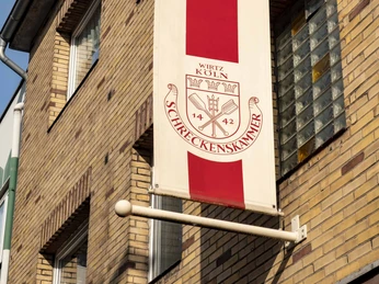 Brewery Schreckenskammer Ein traditionelles Wappenschild des Brauhauses Schreckenskammer hängt an einer Wand aus gepresstem Backstein. Der rote Streifen des Schildes trägt das markante Logo, das neben dem Schriftzug "Schreckenskammer" auch eine detailreiche Illustration eines Wappens zeigt.A traditional emblem shield of the Brauhaus Schreckenskammer mounted on a pressed brick wall. The shield's red stripe prominently features the distinctive logo, which includes the inscription "Schreckenskammer" along with an intricate illustration of a coat of arms.